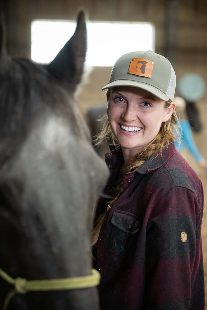 Kat in the barn doorway in a plaid shirt and cap, smiling past the dark horse standing in front of her