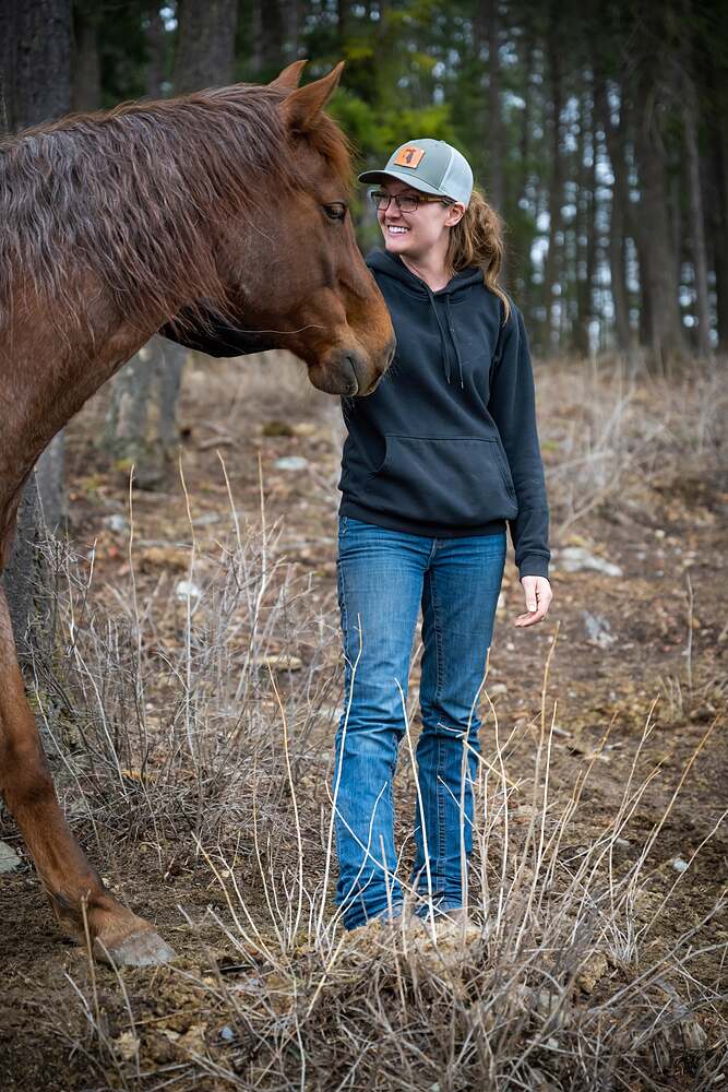 Kat standing in dry spring grass while a loose chestnut horse walks up and reaches its nose toward her