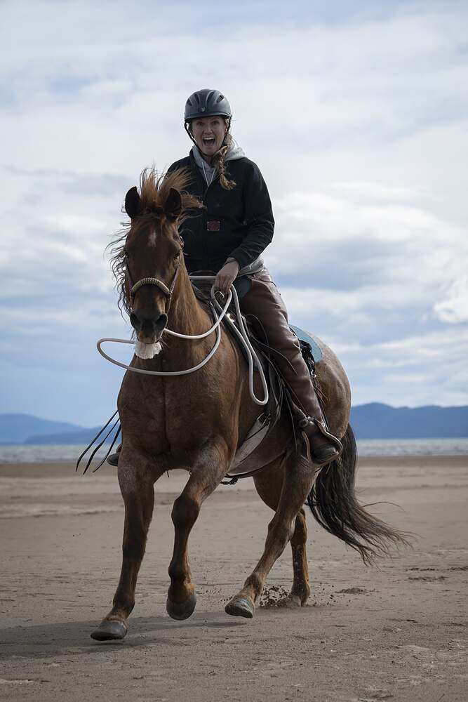 Kat laughing as she rides a chestnut in across the wet sand, the lake behind her