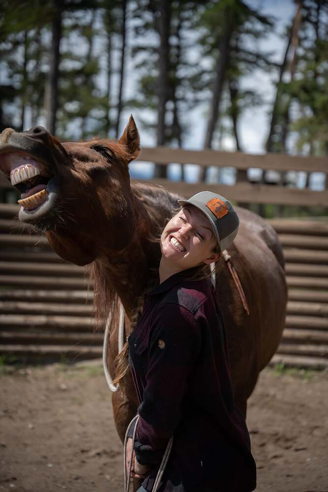 Kat laughing as a chestnut horse pulls a face over her shoulder, teeth bared to the sky